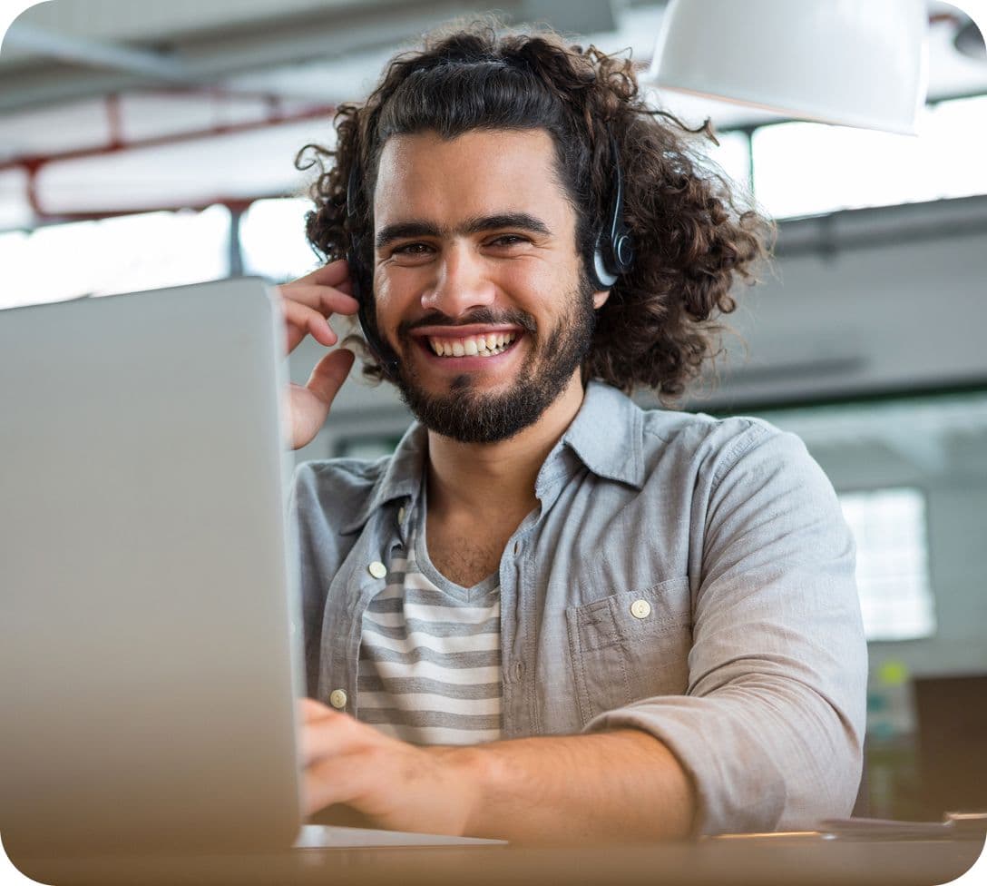 Homem jovem de cabelo cacheado e barba, sorrindo e usando um headset enquanto trabalha em um notebook. Ele transmite uma imagem de profissionalismo e prontidão para oferecer suporte e atendimento.